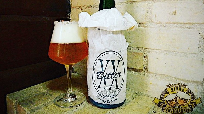 A bottle and glass of De Ranke XX Bitter beer sit on a brick shelf.
