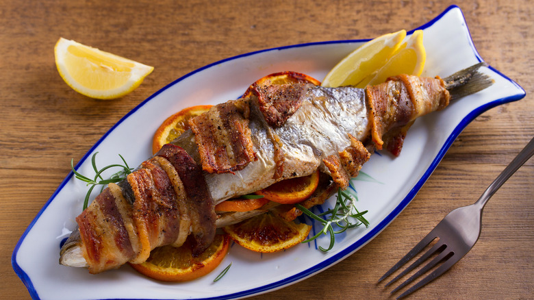 A whole bacon-wrapped fish on a fish-shaped plate with orange and lemon slices