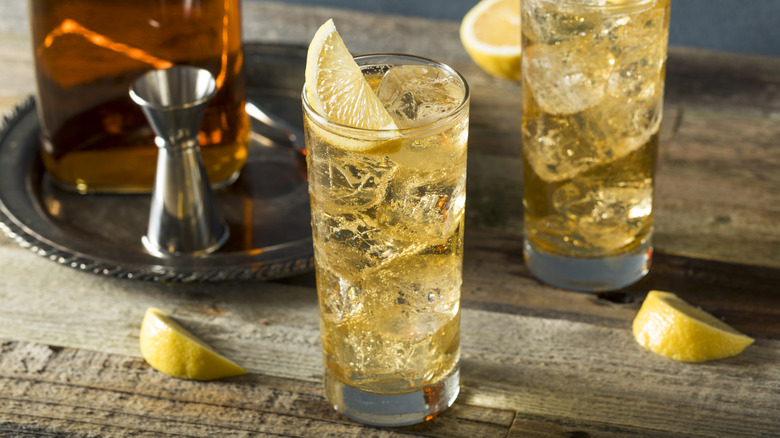 Two highballs on a wooden surface next to lemon wedges, a jigger, and a spirit bottle