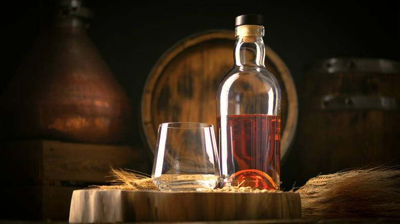 A bottle of whiskey on a barrel next to an empty glass