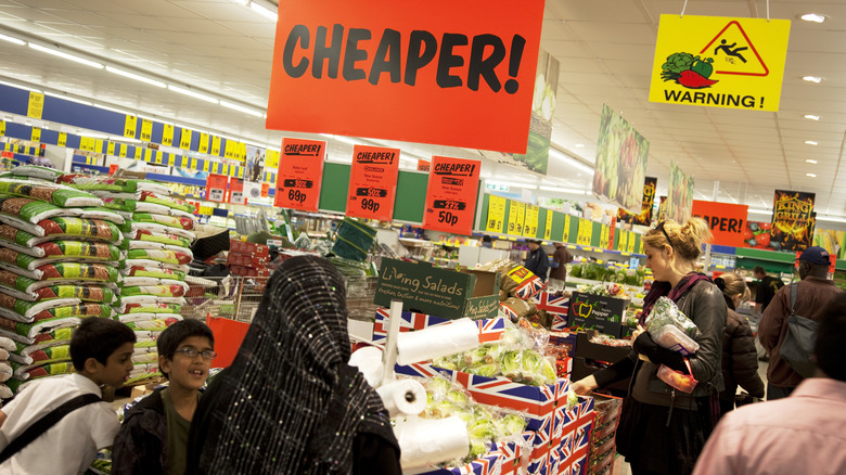 Interior of British Lidl showing people around bins of produce and a red sign with black lettering that reads, "Cheaper!"