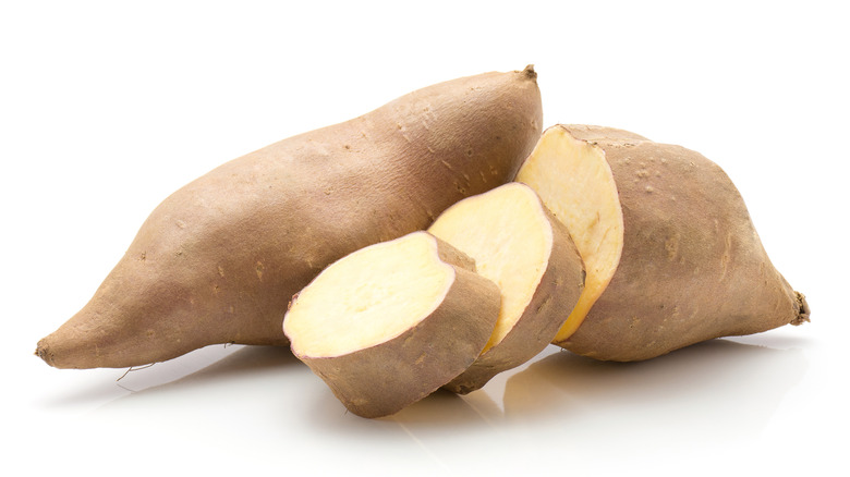 One whole white sweet potato and one sliced white sweet potato on a white surface