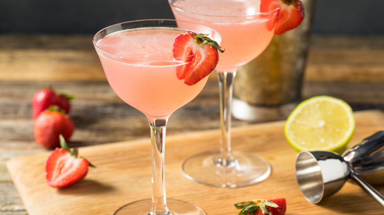 Two strawberry Daiquiris on a wooden surface with fresh strawberries, a jigger, and a lime wedge