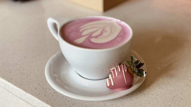 A strawberry-flavored cappuccino in a mug next to a chocolate-covered strawberry