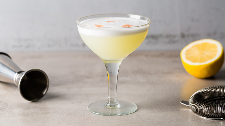 A Pisco Sour cocktail next to a half lemon, jigger, and strainer