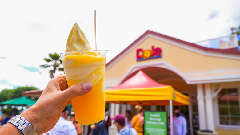 A person's hand holding out a cup of Dole Whip in front of the Dole Plantation.