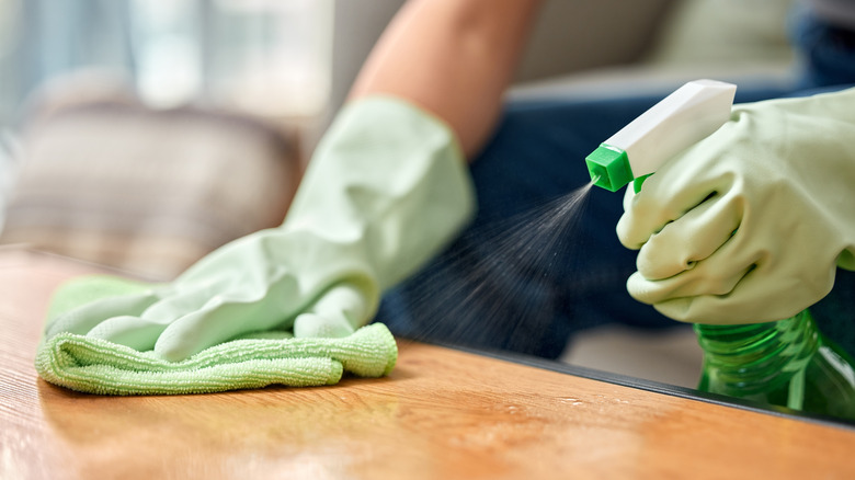 A person with gloves on spraying a wood table and wiping it with a rag