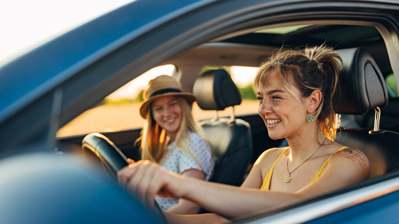 A woman smiling with her hands on a car's steering wheel; another smiling woman sits in the passenger seat