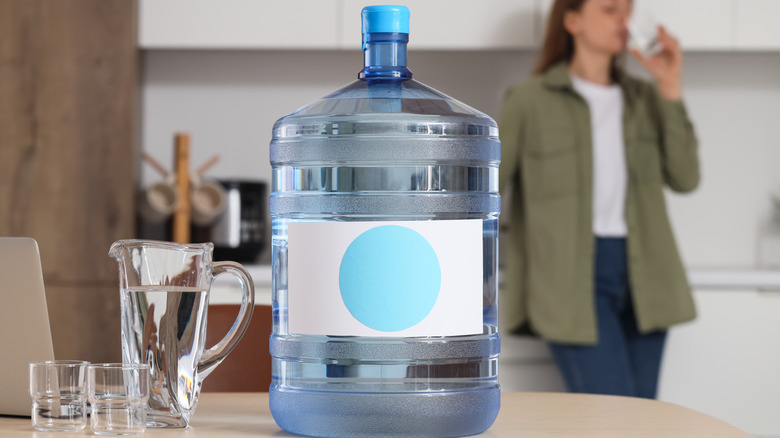 A large jug of water on a kitchen counter; a woman drinks a glass of water in the background