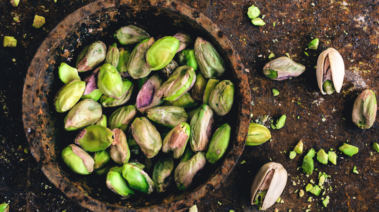 A bowl of shelled pistachios and whole pistachios and crumbs on a brown table