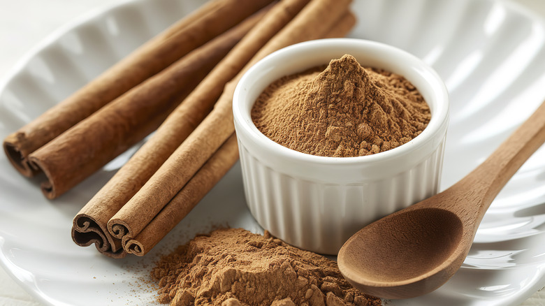 A bowl of cinnamon powder on a plate with a wooden spoon and whole cinnamon sticks