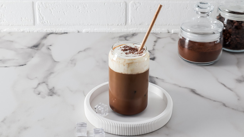 An iced latte on a white plate with cold foam and a brown straw on a marble counter