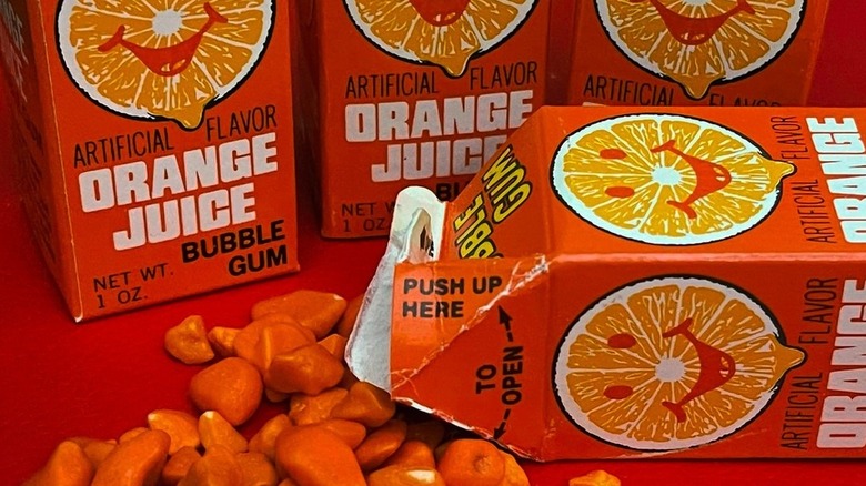 Several packs of Orange Juice Bubble Gum with some spilled out on an orange table