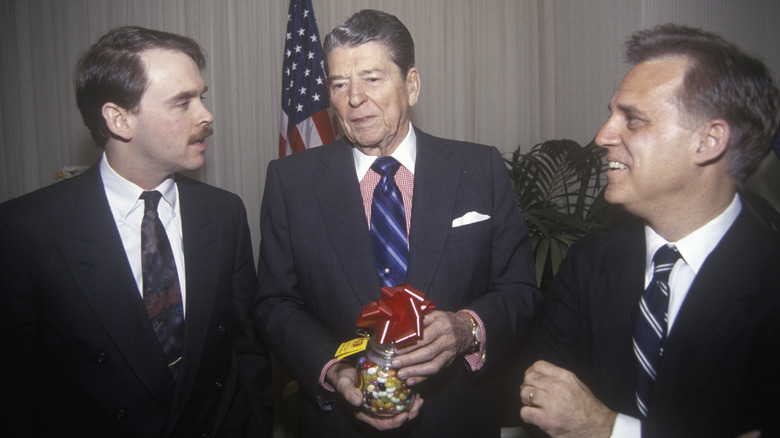 Ronald Reagan stands between two men in suits, holding jar of jelly beans with red bow on top
