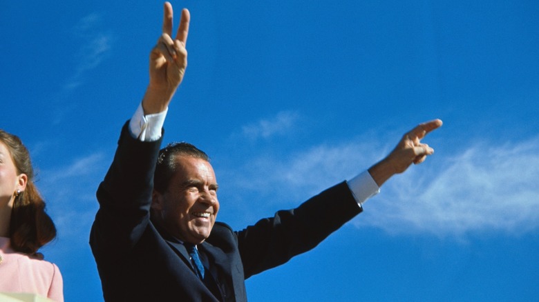 Richard Nixon holds up peace signs while smiling out to crowd, against sky background