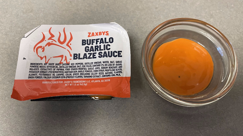 A packet of Buffalo Garlic Blaze Sauce sits on a gray counter next to a dish of bright orange sauce.