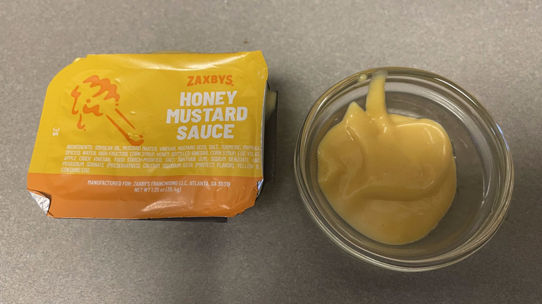 A packet of Honey Mustard Sauce sits on a gray counter next to a dish of yellow sauce.