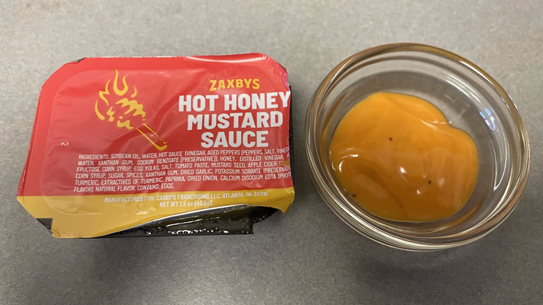 A packet of Hot Honey Mustard Sauce sits on a gray counter next to a dish of light orange sauce.
