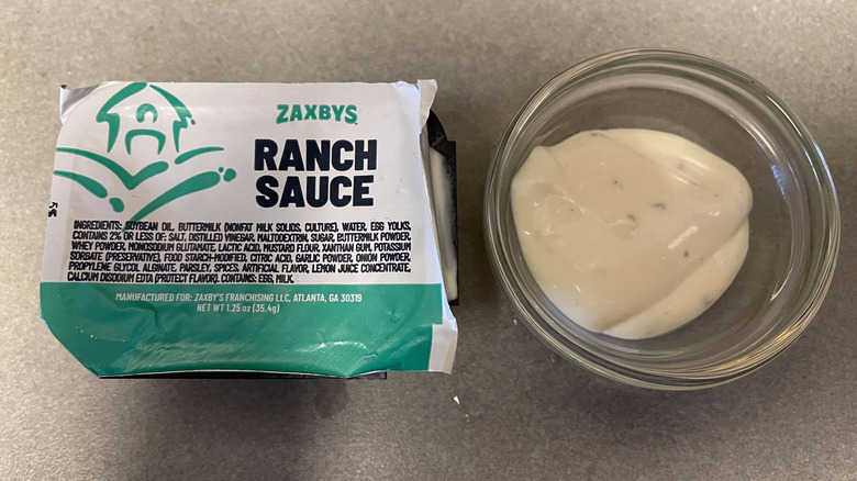 A packet of Ranch Sauce sits on a gray counter next to a dish of white sauce.