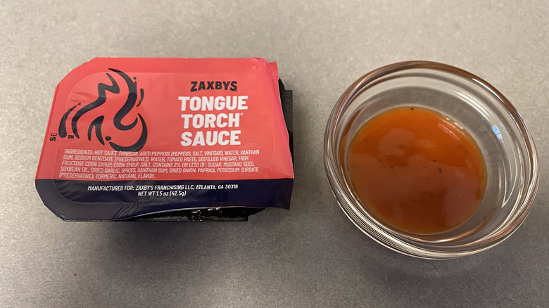 A packet of Tongue Torch Sauce sits on a gray counter next to a dish of dark orange sauce.