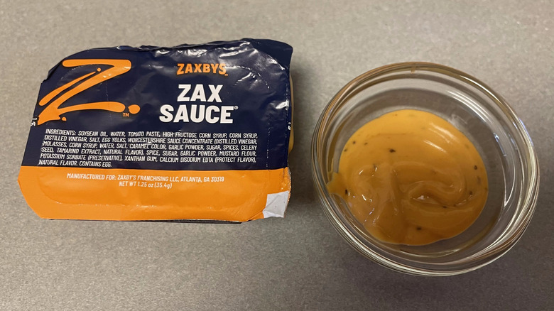 A packet of Zax Sauce sits on a gray counter next to a dish of light orange sauce.