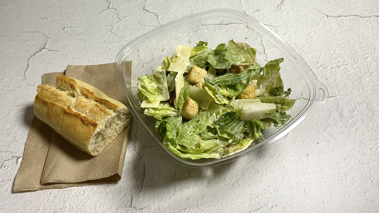 Panera Bread's Caesar Salad in a takeout container next to a piece of French baguette on a paper napkin