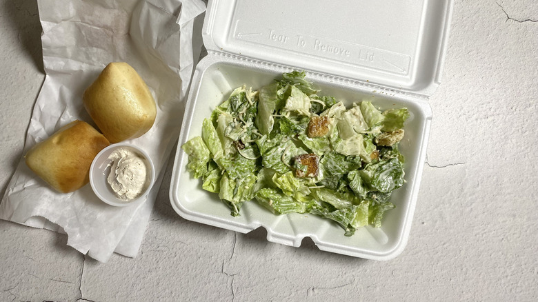 Texas Roadhouse Caesar Salad in a takeout container next to rolls and butter