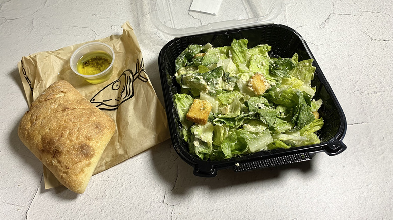 Bonefish Grill's Caesar Salad in a takeout container next to bread and an oil/vinegar mix in a container on top of a paper bag.