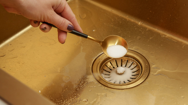 A woman's hand pours a spoonful of baking soda into a sink drain.