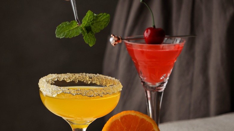 An organe cocktail being garnished with a sprig of mint on tweezers with a red cocktail garnished with a cherry in the back