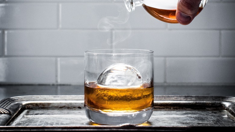 A rocks glass with a large round ball of ice with whisky