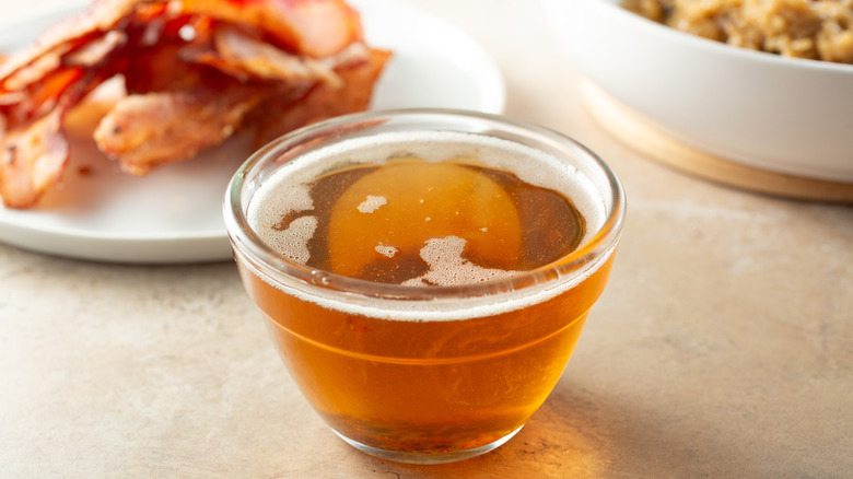 Small glass bowl full of grease with slices of bacon in  the background