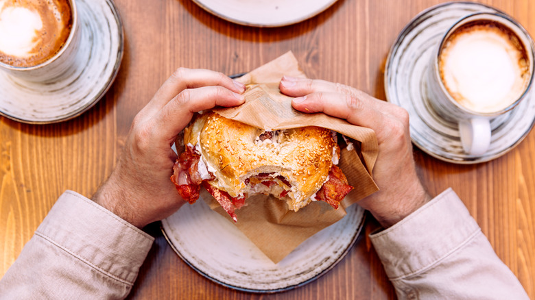 a bagel sandwich with bacon and cream cheese besides cups of coffee