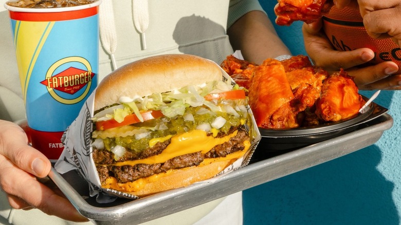 A person holding a tray with a Fatburger cheeseburger, chicken wings, and a drink