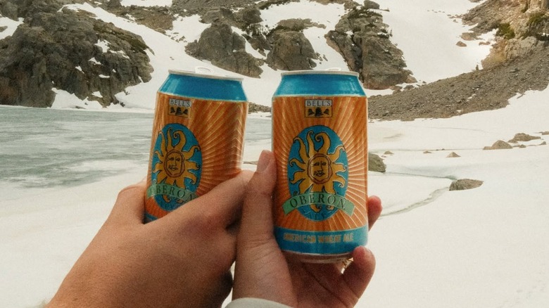 Two hands toasting cans of Bell's Oberon American Wheat Ale on a snowy mountain