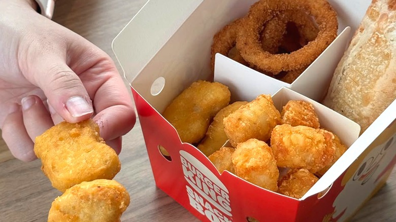 A Snack Box from Burger King with nuggets, tots, a pie, and onion rings next to a person's hand holding a nugget