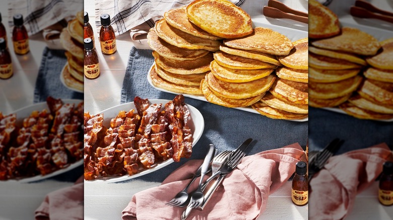 A platter of pancakes next to a plate of bacon strips and syrup bottles