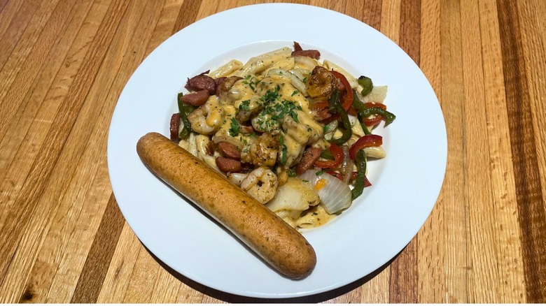 A penne pasta dish with cheese sauce, shrimp, sausage, and peppers sits in a bowl with a breadstick on the side.