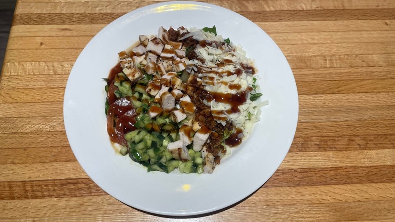 A salad with chicken, cheese, bacon, cucumber, and tomato sits in a bowl on a wood table.