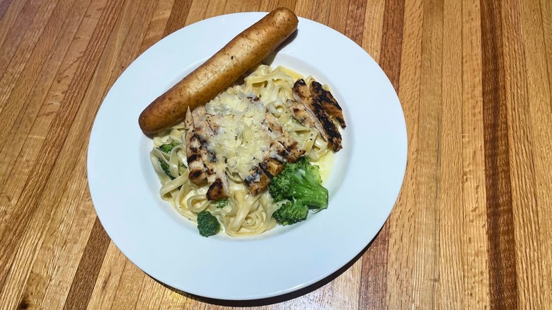 A plate of fettuccine pasta in cream sauce contains chicken, broccoli and parmesan in a bowl with a breadstick on the side.