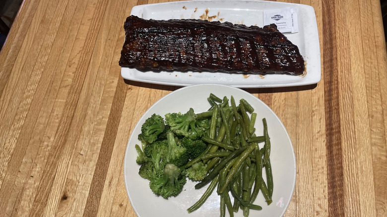 A full rack of ribs site on a plate with a side of broccoli and green beans on another plate beside it.
