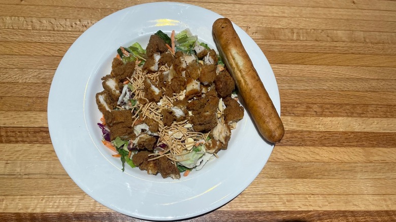 A salad with fried chicken bites, rice noodles, and almonds sits in a bowl with a breadstick on the side.