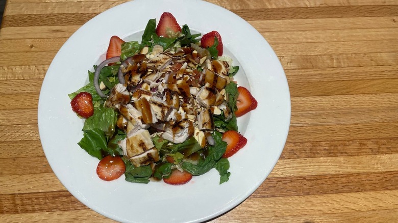 A bed of greens with chicken, onion, strawberries, almonds, and balsamic glaze sits in a bowl on a wood table.