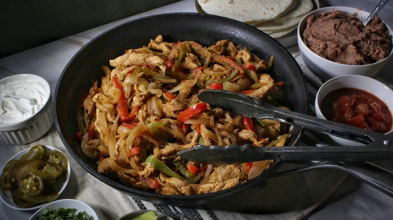 Simple one-pan chicken fajitas in a frying pan with tongs and dishes filled with sides surrounding it.