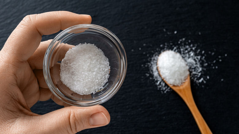 A person's hand is seen holding a tiny bowl of MSG, with a spoon in the background.