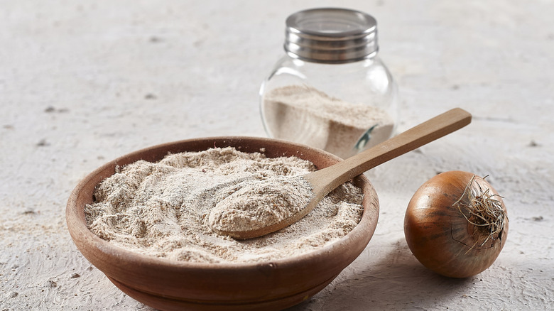 A wooden bowl full of onion powder joined by an onion and a seasoning shaker.