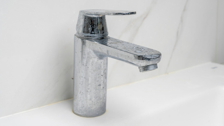 Chrome sink faucet covered with scale on a white sink and in front of a white marble wall