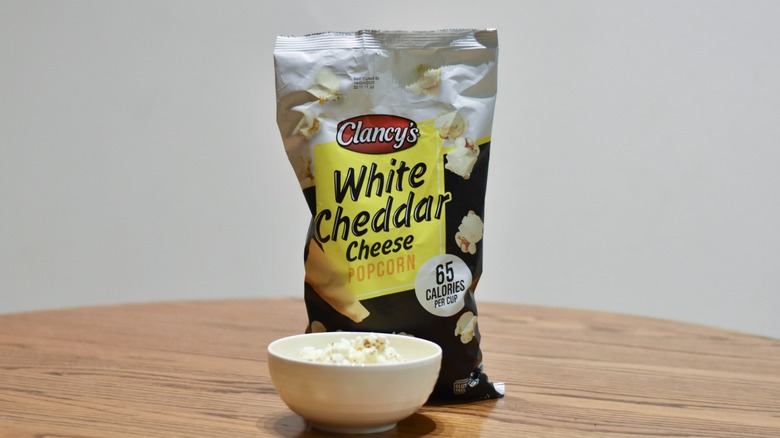A bag of Clancy' white cheddar cheese popcorn next to a white bowl of popcorn