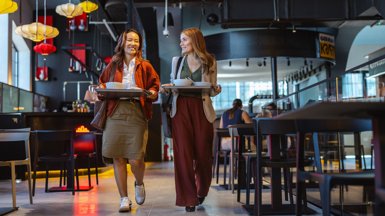 Two people carrying trays of food in a mall food court