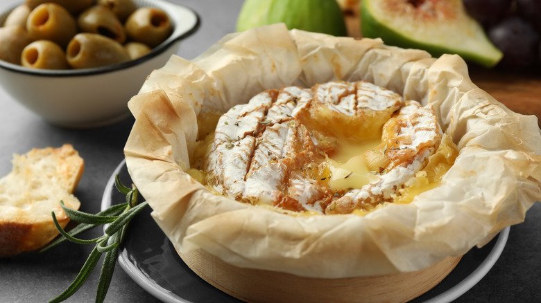 Baked brie in parchment paper on a table with bread, olives, and fruit